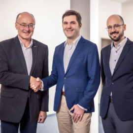 Drei Personen stehen in einem Innenraum, zwei davon geben sich die Hand; alle tragen Businesskleidung, links steht eine Pflanze, rechts ein Banner.