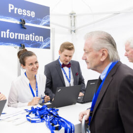Mehrere Personen stehen um einen Tisch mit Laptops und tragen blaue Namensbänder. Über dem Tisch hängt ein Schild mit der Aufschrift „Presse Information“. Die Szene spielt sich an einem Informationsstand ab, vermutlich im Rahmen einer Veranstaltung oder Konferenz.
