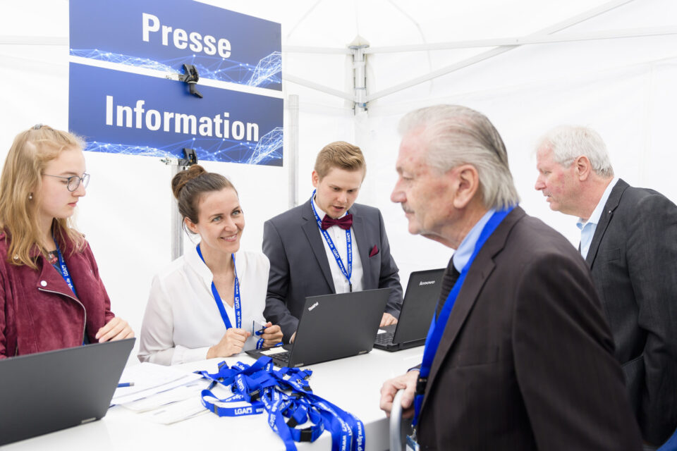Mehrere Personen stehen um einen Tisch mit Laptops und tragen blaue Namensbänder. Über dem Tisch hängt ein Schild mit der Aufschrift „Presse Information“. Die Szene spielt sich an einem Informationsstand ab, vermutlich im Rahmen einer Veranstaltung oder Konferenz.