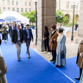 Mehrere Personen gehen über einen blauen Teppich unter einem Torbogen hindurch. Einige tragen moderne Anzüge, andere historische Kostüme. Im Hintergrund befinden sich ein Zelt mit weiteren Menschen sowie Gebäude mit großen Fenstern und etwas Grünfläche.
