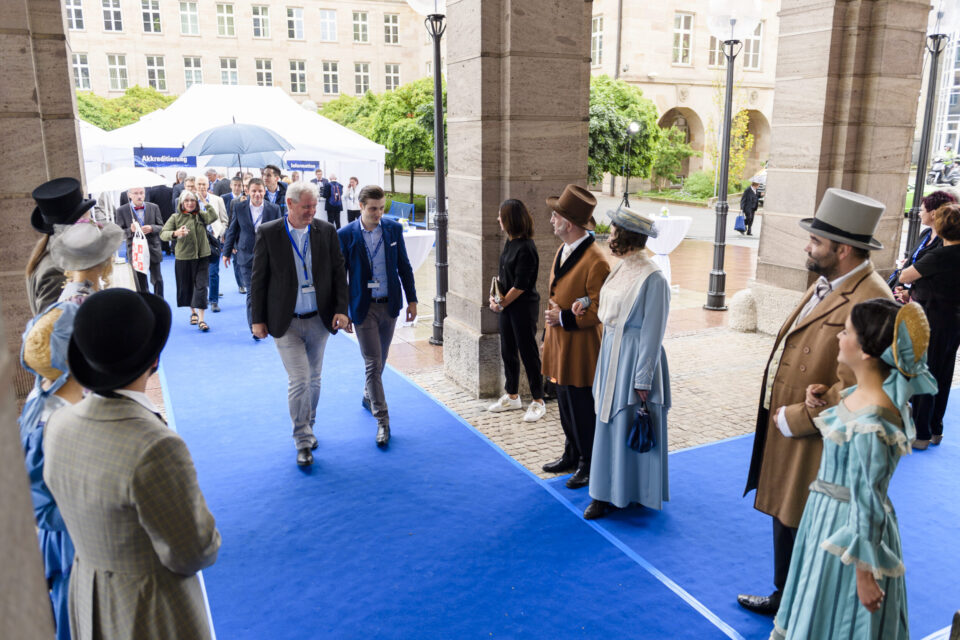 Mehrere Personen gehen über einen blauen Teppich unter einem Torbogen hindurch. Einige tragen moderne Anzüge, andere historische Kostüme. Im Hintergrund befinden sich ein Zelt mit weiteren Menschen sowie Gebäude mit großen Fenstern und etwas Grünfläche.