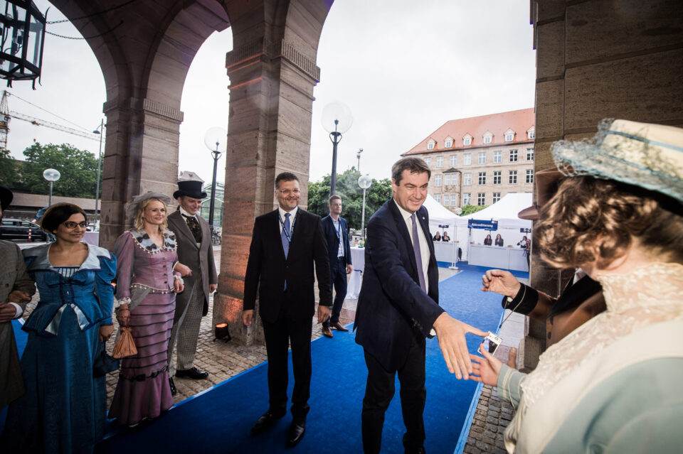 Das Bild zeigt eine Gruppe von Menschen auf einem blauen Teppich unter einem Torbogen, darunter Personen in historischer Kleidung und zwei Männer in modernen Anzügen. Einer der Männer in Anzug schüttelt einer Person in historischer Kleidung die Hand. Im Hintergrund sind ein Gebäude mit vielen Fenstern sowie Zelte oder Stände zu sehen, was auf eine Veranstaltung mit historischem Bezug hinweist.