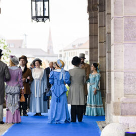 Das Bild zeigt eine Gruppe von Menschen in historischen Kostümen, die auf einem blauen Teppich unter einem Torbogen stehen. Die Kleidung erinnert an das 19. Jahrhundert, mit Zylindern, langen Kleidern mit Reifröcken und Anzügen mit Gehrock. Die Szene wirkt wie eine Veranstaltung im Freien an einem geschichtlich oder kulturell bedeutsamen Ort.
