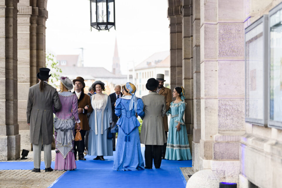 Das Bild zeigt eine Gruppe von Menschen in historischen Kostümen, die auf einem blauen Teppich unter einem Torbogen stehen. Die Kleidung erinnert an das 19. Jahrhundert, mit Zylindern, langen Kleidern mit Reifröcken und Anzügen mit Gehrock. Die Szene wirkt wie eine Veranstaltung im Freien an einem geschichtlich oder kulturell bedeutsamen Ort.