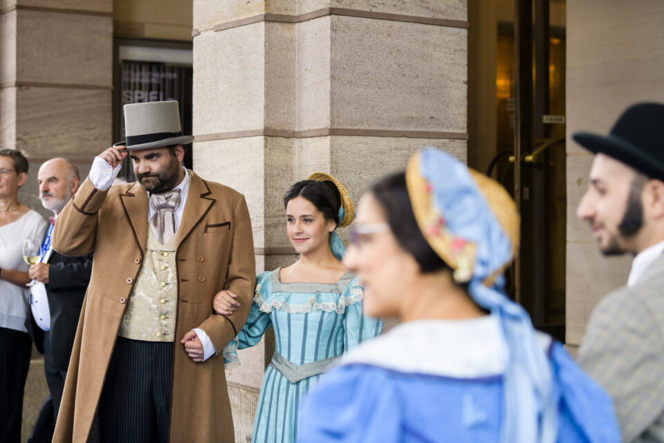 Das Bild zeigt eine Gruppe von Menschen in historischen Kostümen vor einem Gebäude mit steinernen Säulen. Im Mittelpunkt steht ein Mann mit braunem Mantel, beigem Westenanzug und Zylinder, der den Arm einer Frau in einem blauen Kleid mit Spitzenbesatz hält. Weitere Personen in historischer Kleidung sind im Hintergrund zu sehen.