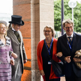 Das Bild zeigt eine Gruppe von Menschen neben einer steinernen Säule. Zwei Personen auf der linken Seite tragen historische Kleidung, darunter ein lila Kleid mit Hut und ein Anzug mit Zylinder. Auf der rechten Seite stehen zwei Personen in moderner Kleidung, eine mit roter Jacke und die andere im Anzug mit einem Mantel über dem Arm.