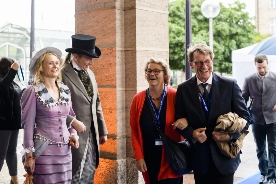 Das Bild zeigt eine Gruppe von Menschen neben einer steinernen Säule. Zwei Personen auf der linken Seite tragen historische Kleidung, darunter ein lila Kleid mit Hut und ein Anzug mit Zylinder. Auf der rechten Seite stehen zwei Personen in moderner Kleidung, eine mit roter Jacke und die andere im Anzug mit einem Mantel über dem Arm.