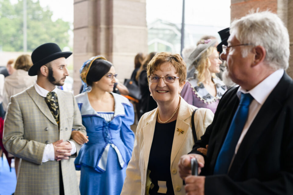 Das Bild zeigt eine Gruppe von Menschen in historischer oder nostalgischer Kleidung. Im Vordergrund sind fünf Personen zu sehen, darunter eine Person mit Melone, hellem Anzug und weißen Handschuhen sowie eine weitere in einem blauen Kleid mit weißen Akzenten und Kopftuch. Weitere Personen tragen elegante Kleidung mit Accessoires wie Perlen, Broschen oder einem Gehstock. Im Hintergrund befinden sich weitere Menschen in ähnlicher historischer Kleidung, was auf eine Veranstaltung mit historischem Modethema hinweist.