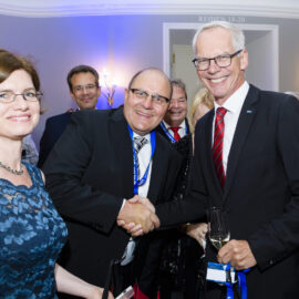 Das Bild zeigt eine Gruppe von Menschen bei einer formellen Veranstaltung. Zwei Männer in der Mitte geben sich die Hand, beide tragen Anzüge, einer hält ein Glas Wein. Links steht eine Frau in einem blauen Kleid, im Hintergrund sind weitere Personen sowie Wandlampen und ein Schild über einer Tür mit der Aufschrift „REIHEN 18–20“ zu sehen.