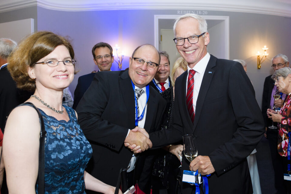 Das Bild zeigt eine Gruppe von Menschen bei einer formellen Veranstaltung. Zwei Männer in der Mitte geben sich die Hand, beide tragen Anzüge, einer hält ein Glas Wein. Links steht eine Frau in einem blauen Kleid, im Hintergrund sind weitere Personen sowie Wandlampen und ein Schild über einer Tür mit der Aufschrift „REIHEN 18–20“ zu sehen.