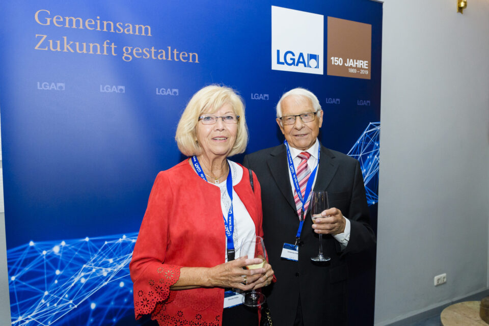 Das Bild zeigt zwei Personen in formeller Kleidung, die vor einem blauen Hintergrund mit der Aufschrift „Gemeinsam Zukunft gestalten“. Beide tragen Namensschilder an einem Band um den Hals und Weingläser.