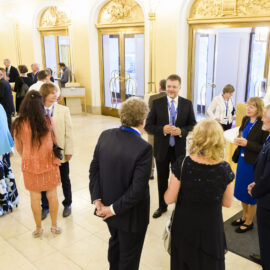 Das Bild zeigt eine Gruppe von Menschen bei einer formellen Veranstaltung. Die Personen tragen überwiegend Geschäfts- oder festliche Kleidung und Namensschilder an Bändern um den Hals. Der Raum ist elegant gestaltet mit verzierten Türen und großen Glasflächen, rechts befindet sich ein Empfangstisch, an dem zwei Personen im Gespräch sind.