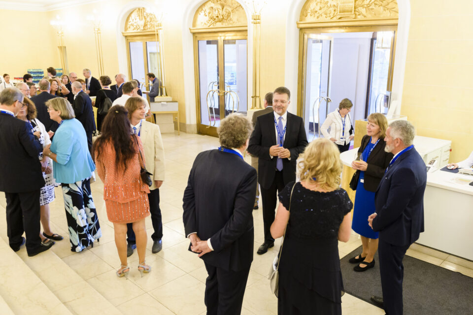 Das Bild zeigt eine Gruppe von Menschen bei einer formellen Veranstaltung. Die Personen tragen überwiegend Geschäfts- oder festliche Kleidung und Namensschilder an Bändern um den Hals. Der Raum ist elegant gestaltet mit verzierten Türen und großen Glasflächen, rechts befindet sich ein Empfangstisch, an dem zwei Personen im Gespräch sind.
