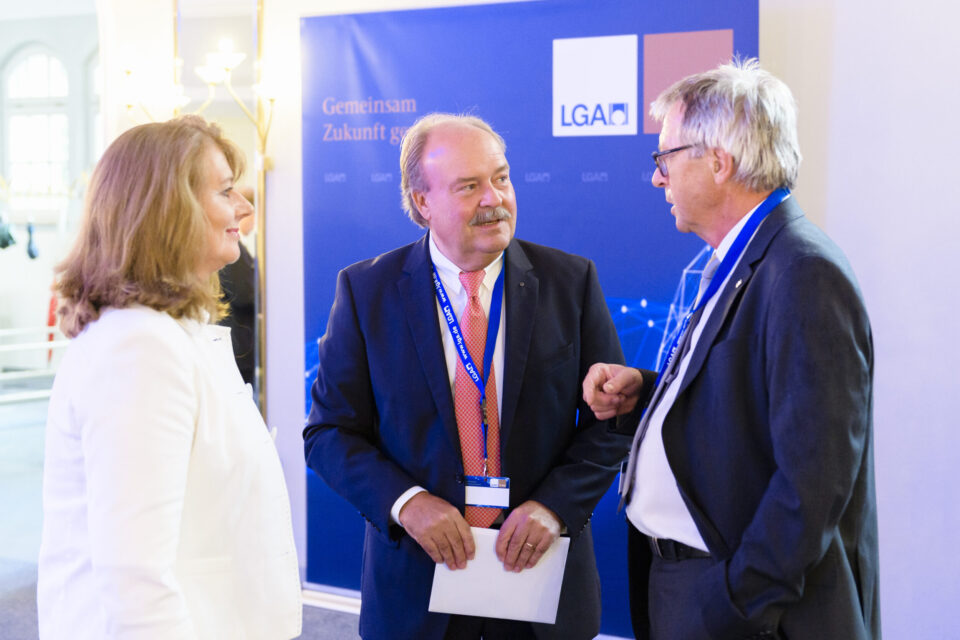 Das Bild zeigt drei Personen im Gespräch bei einer professionellen oder formellen Veranstaltung. Sie stehen vor einem blauen Hintergrund mit der Aufschrift „Gemeinsam Zukunft gestalten“ und dem Logo „LGA“. Alle tragen Businesskleidung und Namensschilder.