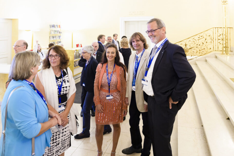 Das Bild zeigt eine Gruppe von Menschen, die sich in einem formellen oder professionellen Rahmen unterhalten. Alle tragen Namensschilder an Bändern, was auf eine Veranstaltung hinweist. Im Hintergrund sind eine Treppe mit verzierten Geländern, Regale mit Büchern oder Materialien sowie Türen zu weiteren Räumen zu sehen.