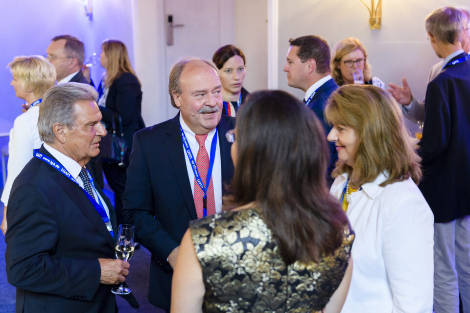 Mehrere Personen stehen in einem Innenraum bei einer formellen Veranstaltung oder Konferenz. Sie tragen Businesskleidung und Namensschilder an blauen Bändern, unterhalten sich und halten teilweise Getränke in der Hand.