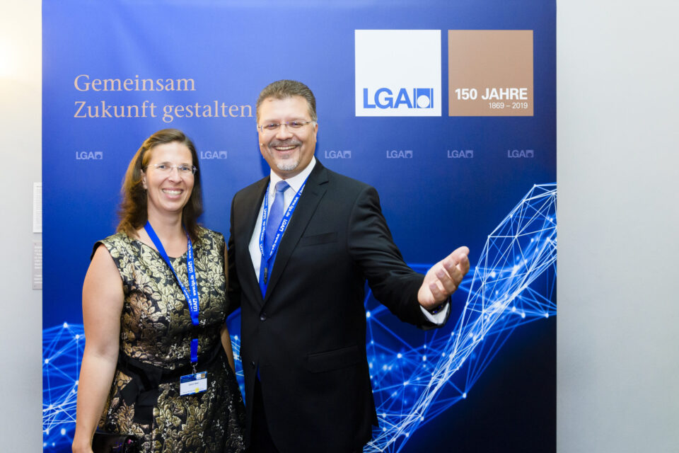 Zwei Personen stehen vor einem blauen Hintergrund mit Logos und einem Jubiläumshinweis. Beide tragen formelle Kleidung und Namensschilder an Bändern um den Hals.