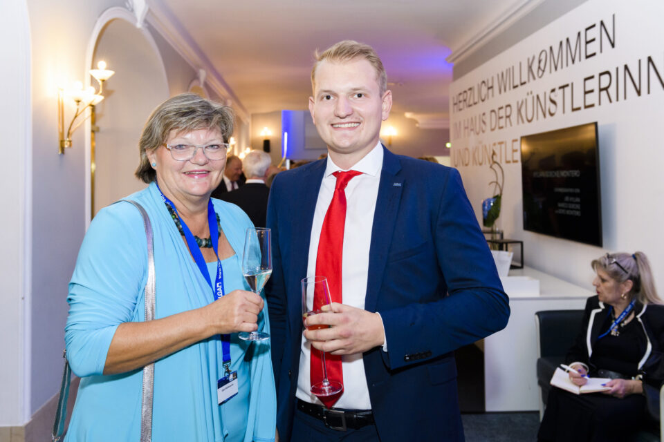 Zwei Personen stehen in einem hell erleuchteten Raum und halten Getränke in der Hand. Die linke Person trägt ein hellblaues Outfit, die rechte einen dunkelblauen Anzug mit weißem Hemd und roter Krawatte. Im Hintergrund sind weitere Menschen, Möbel sowie ein Bildschirm und ein Willkommensschild an der Wand zu sehen.