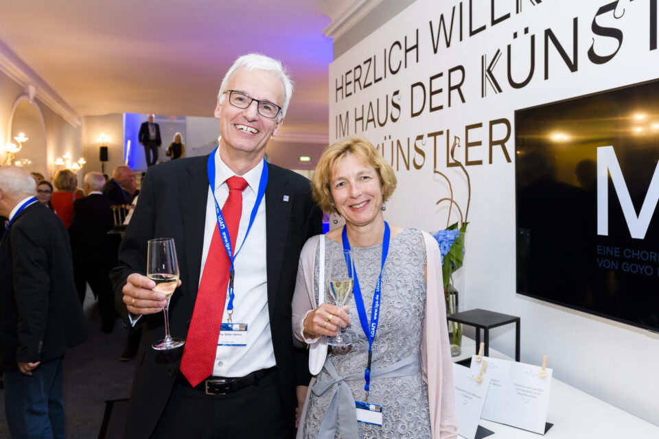 Zwei Personen stehen nebeneinander bei einer Veranstaltung und halten jeweils ein Glas Wein in der Hand. Beide tragen Namensschilder an blauen Bändern. Im Hintergrund ist eine Wand mit einem Willkommensschriftzug.