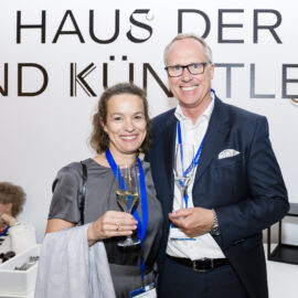 Drei Personen befinden sich in einem Raum mit einer weißen Wand, auf der ein Schriftzug zu sehen ist. Zwei Personen stehen und halten Gläser mit Sekt, eine weitere Person sitzt an einem Tisch mit einigen Gegenständen darauf. Im Hintergrund sind Blumen und kleine Tische als Dekoration zu erkennen.