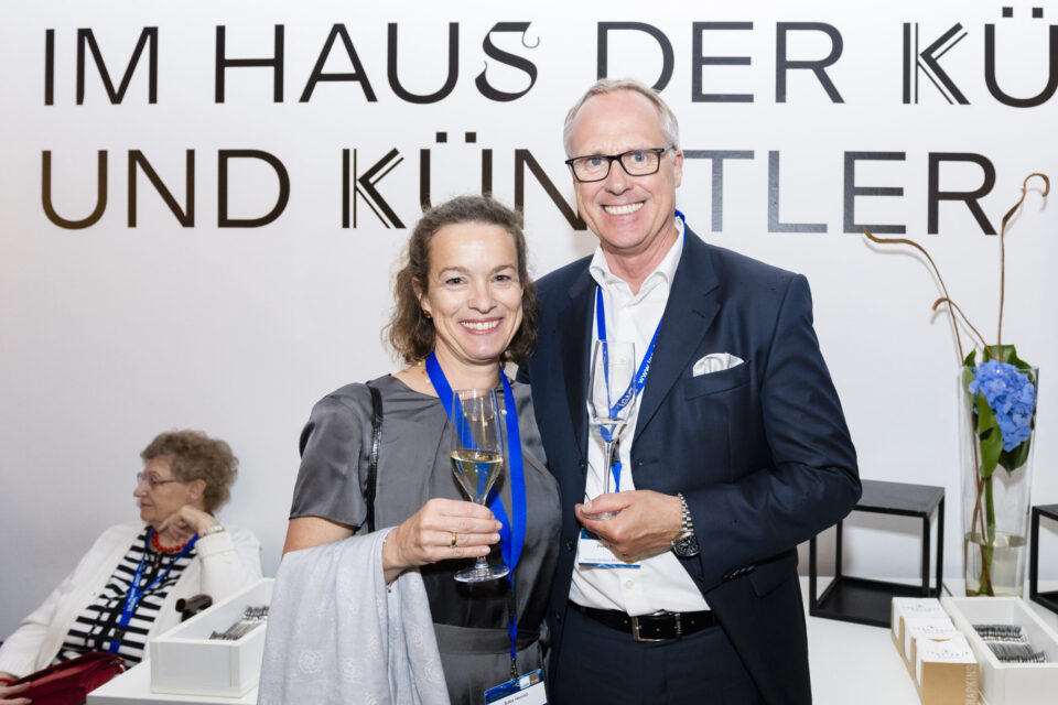 Drei Personen befinden sich in einem Raum mit einer weißen Wand, auf der ein Schriftzug zu sehen ist. Zwei Personen stehen und halten Gläser mit Sekt, eine weitere Person sitzt an einem Tisch mit einigen Gegenständen darauf. Im Hintergrund sind Blumen und kleine Tische als Dekoration zu erkennen.