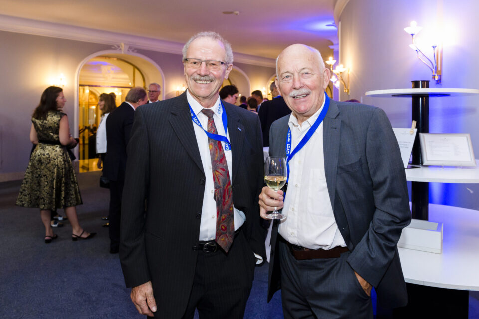 Bei einer gesellschaftlichen Veranstaltung stehen zwei Personen im Vordergrund, beide in Anzügen und mit blauen Namensbändern. Eine der Personen hält ein Glas Weißwein. Im Hintergrund unterhalten sich weitere Gäste in einem elegant gestalteten Innenraum mit weicher Beleuchtung und dekorativen Elementen wie Wandlampen und Rundbögen.