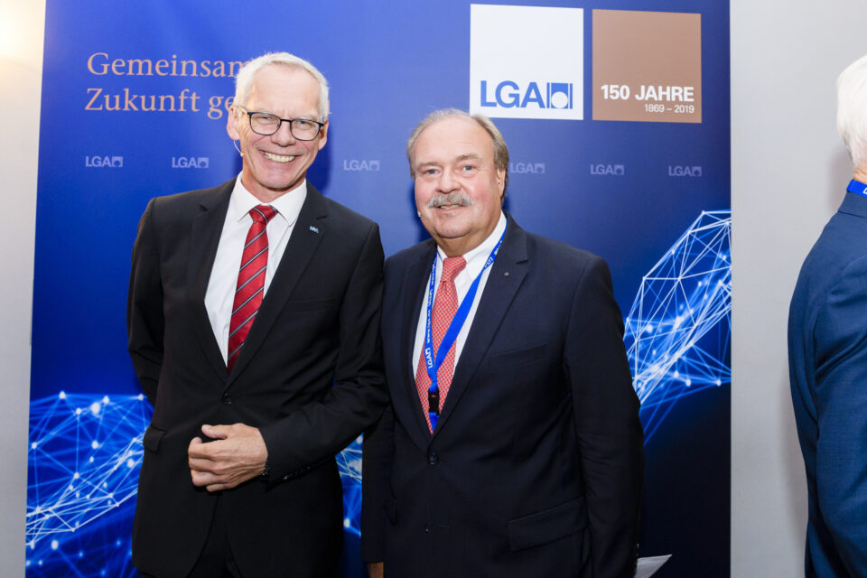 Zwei Personen in Businesskleidung stehen vor einem blauen Hintergrund mit grafischen Elementen aus Linien und Knotenpunkten. Der Hintergrund zeigt Logos und einen Jubiläumshinweis.