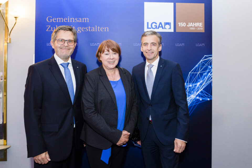 Drei Personen stehen vor einem blauen Hintergrund mit Logos und einem Jubiläumshinweis. Der Hintergrund zeigt den Schriftzug einer gemeinsamen Zukunftsgestaltung.