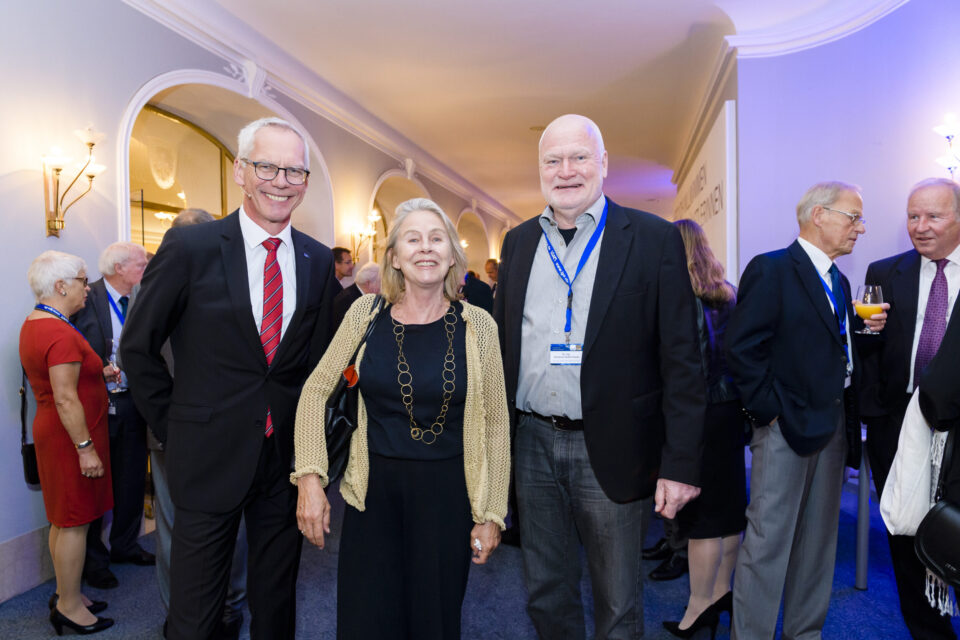 Drei Personen stehen im Vordergrund einer Veranstaltung in einem eleganten Raum mit stilvollen Leuchten und Rundbögen. Weitere Gäste sind im Hintergrund zu sehen, einige tragen Namensschilder an Bändern. Die Kleidung der Anwesenden ist formell, was auf ein Event hinweist.