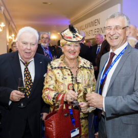 Mehrere Personen in formeller Kleidung stehen bei einer gesellschaftlichen oder beruflichen Veranstaltung zusammen. Sie tragen Namensschilder an blauen Bändern und halten Gläser mit Getränken. Im Hintergrund ist ein Schild mit einem teilweise sichtbaren Willkommensgruß zu erkennen.