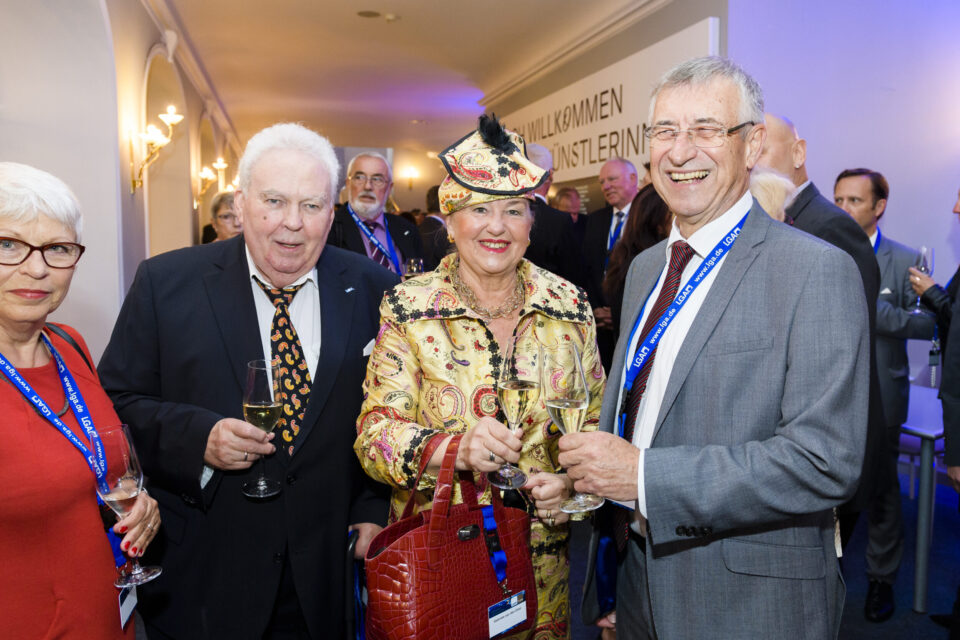 Mehrere Personen in formeller Kleidung stehen bei einer gesellschaftlichen oder beruflichen Veranstaltung zusammen. Sie tragen Namensschilder an blauen Bändern und halten Gläser mit Getränken. Im Hintergrund ist ein Schild mit einem teilweise sichtbaren Willkommensgruß zu erkennen.