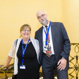 Zwei Personen stehen nebeneinander in einem gut beleuchteten Innenraum. Beide tragen blaue Namensbänder, die linke Person ein schwarzes Outfit, die rechte einen dunklen Anzug mit gemusterter Krawatte. Im Hintergrund sind helle Wände und verzierte Metallgeländer zu sehen.