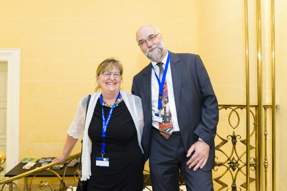 Zwei Personen stehen nebeneinander in einem gut beleuchteten Innenraum. Beide tragen blaue Namensbänder, die linke Person ein schwarzes Outfit, die rechte einen dunklen Anzug mit gemusterter Krawatte. Im Hintergrund sind helle Wände und verzierte Metallgeländer zu sehen.