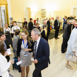 Mehrere Personen befinden sich in einem hell beleuchteten Innenraum mit Marmorboden bei einer formellen Veranstaltung oder Konferenz. Einige unterhalten sich, tragen Namensschilder an Bändern oder halten Unterlagen in der Hand. Im Hintergrund ist ein Empfangstresen mit Personal sowie Regale mit Broschüren oder Büchern zu sehen.