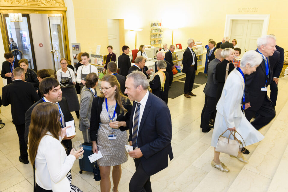 Mehrere Personen befinden sich in einem hell beleuchteten Innenraum mit Marmorboden bei einer formellen Veranstaltung oder Konferenz. Einige unterhalten sich, tragen Namensschilder an Bändern oder halten Unterlagen in der Hand. Im Hintergrund ist ein Empfangstresen mit Personal sowie Regale mit Broschüren oder Büchern zu sehen.