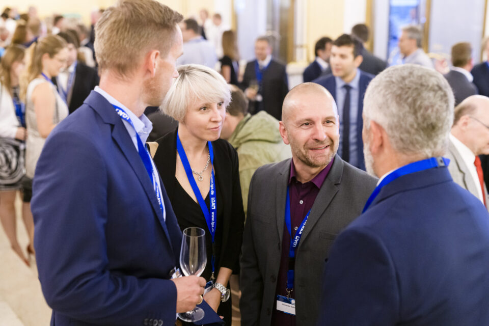 Mehrere Personen befinden sich bei einer beruflichen oder Netzwerkveranstaltung und unterhalten sich miteinander. Sie tragen Namensschilder an blauen Bändern, eine Person hält ein leeres Weinglas. Im Hintergrund sind weitere Gäste in Gespräche vertieft, was auf eine gesellige Atmosphäre hinweist.