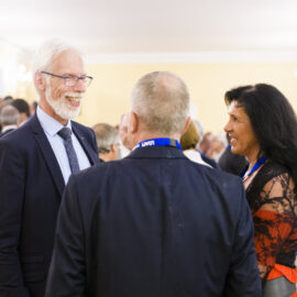 Drei Personen stehen im Vordergrund einer Veranstaltung in einem elegant gestalteten Innenraum mit heller Beleuchtung und Kronleuchtern. Sie unterhalten sich, während im Hintergrund weitere Gäste in Gespräche vertieft sind. Einige Anwesende tragen Namensschilder oder Bänder, was auf eine Konferenz oder ein Netzwerktreffen hinweist.