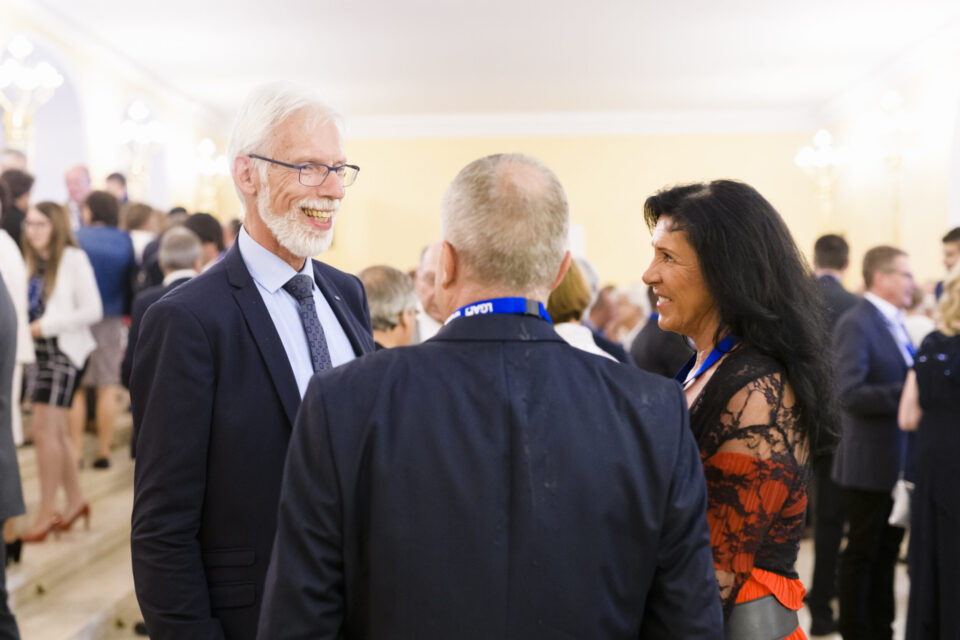 Drei Personen stehen im Vordergrund einer Veranstaltung in einem elegant gestalteten Innenraum mit heller Beleuchtung und Kronleuchtern. Sie unterhalten sich, während im Hintergrund weitere Gäste in Gespräche vertieft sind. Einige Anwesende tragen Namensschilder oder Bänder, was auf eine Konferenz oder ein Netzwerktreffen hinweist.