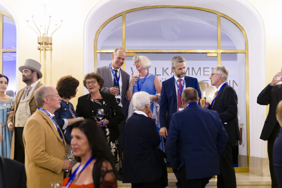 Mehrere Personen in formeller Kleidung stehen in einem eleganten Innenraum mit warmem Licht und dekorativen Elementen wie Kronleuchtern. Im Hintergrund ist ein großer Rundbogen mit goldenen Verzierungen und ein teilweise sichtbares Willkommensschild zu sehen. Einige Gäste halten Getränke und unterhalten sich.