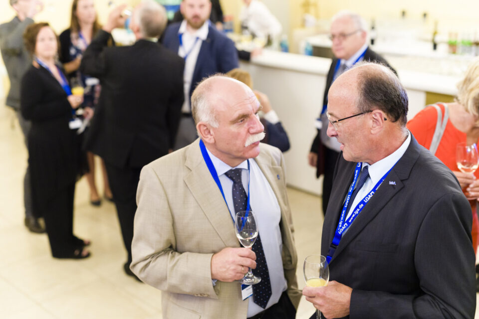 Mehrere Personen befinden sich bei einer gesellschaftlichen oder beruflichen Veranstaltung und tragen Businesskleidung sowie Namensschilder an blauen Bändern. Einige Gäste halten Getränke in der Hand und unterhalten sich.