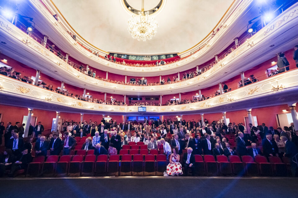 Das Bild zeigt das Innere eines prunkvollen Theaters oder Opernhauses mit mehreren Rängen und einem großen Kronleuchter an der Decke. Die Zuschauer sitzen in roten Stühlen, einige stehen oder gehen umher, was auf eine Pause oder den Beginn der Veranstaltung hindeutet. Die Architektur ist reich verziert mit weißen und goldenen Elementen.