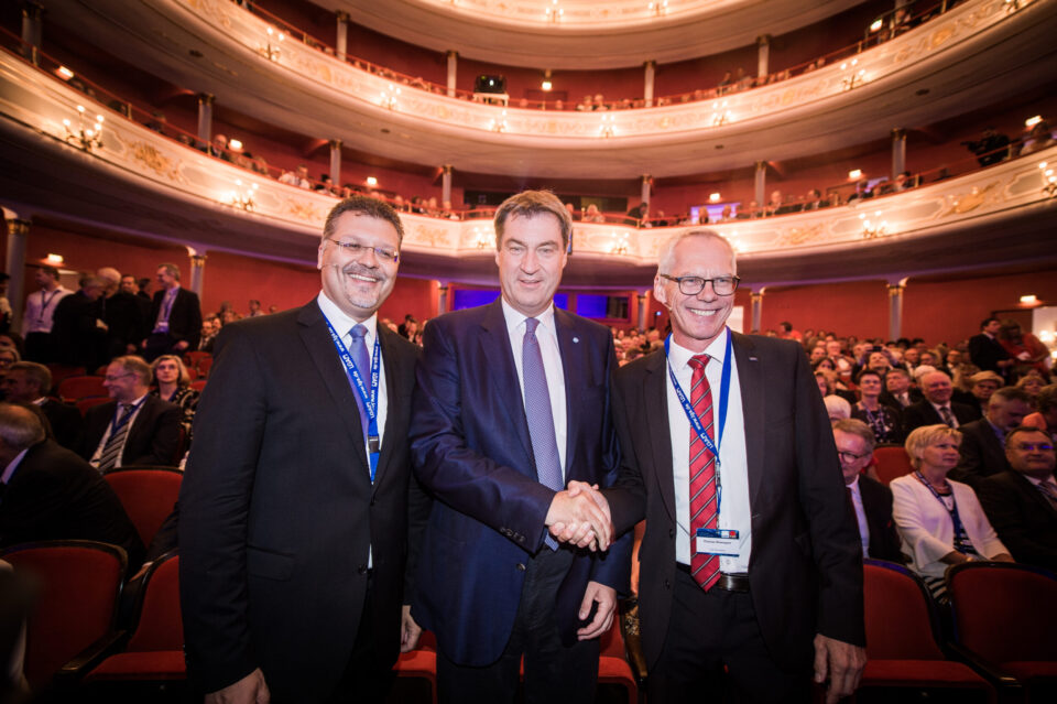 Das Bild zeigt drei Personen in formeller Kleidung, die sich im Vordergrund eines großen, prunkvollen Theaters oder Auditoriums die Hand geben. Zwei der Personen tragen Namensschilder an einem Band um den Hals. Im Hintergrund sind mehrere Ränge mit zahlreichen Zuschauerinnen und Zuschauern zu sehen, was auf eine gut besuchte Veranstaltung hinweist.