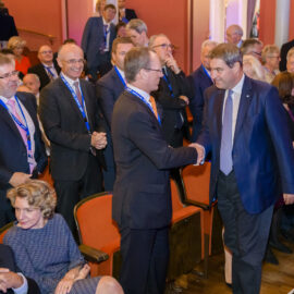 Mehrere Personen befinden sich in einem formellen Veranstaltungsraum, viele tragen Anzüge und Krawatten. Zwei Männer im Vordergrund geben sich die Hand, während andere Gäste sitzen oder sich unterhalten. Im Hintergrund ist eine rote Wand zu sehen.