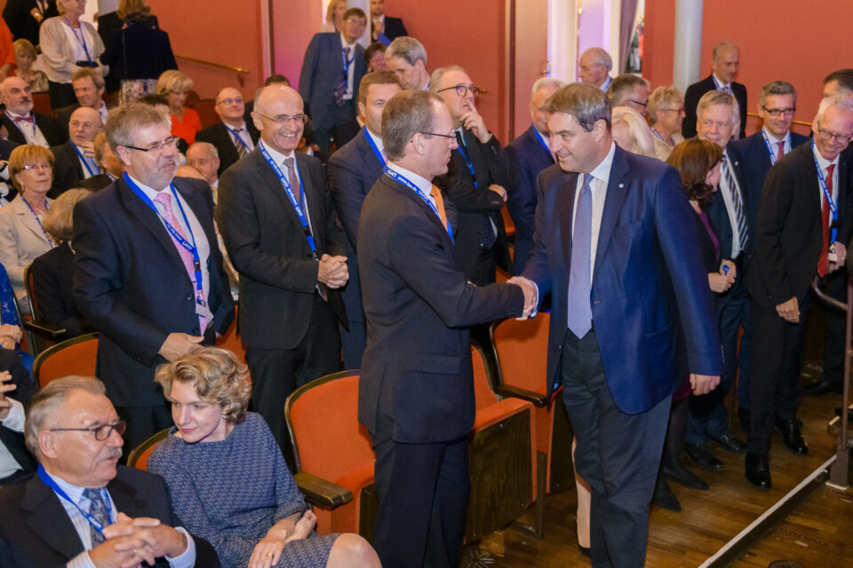 Mehrere Personen befinden sich in einem formellen Veranstaltungsraum, viele tragen Anzüge und Krawatten. Zwei Männer im Vordergrund geben sich die Hand, während andere Gäste sitzen oder sich unterhalten. Im Hintergrund ist eine rote Wand zu sehen.