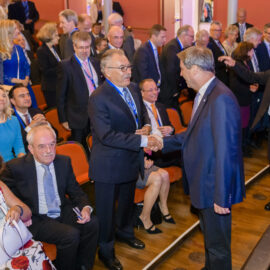 Mehrere Personen in formeller Kleidung befinden sich in einem Saal mit orangefarbenen Stühlen und Holzfußboden. Einige stehen und geben sich die Hand, während andere sitzen. Die Umgebung deutet auf einen Konferenz- oder Veranstaltungsraum hin.