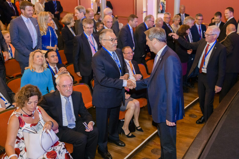 Mehrere Personen in formeller Kleidung befinden sich in einem Saal mit orangefarbenen Stühlen und Holzfußboden. Einige stehen und geben sich die Hand, während andere sitzen. Die Umgebung deutet auf einen Konferenz- oder Veranstaltungsraum hin.