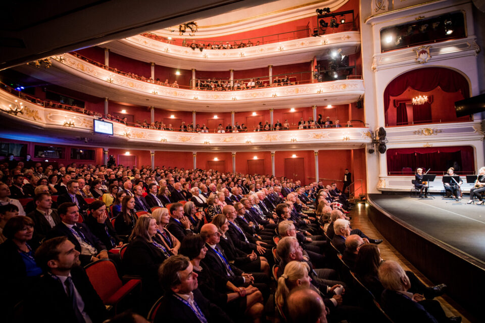 Das Bild zeigt ein prunkvolles Theater mit roten Wänden und goldenen Verzierungen, in dem zahlreiche Menschen in mehreren Sitzreihen und auf Balkonen Platz genommen haben. Auf der rechten Seite befindet sich eine Bühne, auf der einige Musiker auftreten. Das Publikum verfolgt die Darbietung aufmerksam.