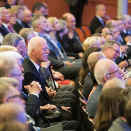 Viele Personen sitzen in einem Auditorium oder Konferenzsaal in Reihen angeordneten Stühlen. Sie tragen formelle Kleidung und wirken aufmerksam, als würden sie einer Veranstaltung wie einer Konferenz oder einem Seminar folgen.