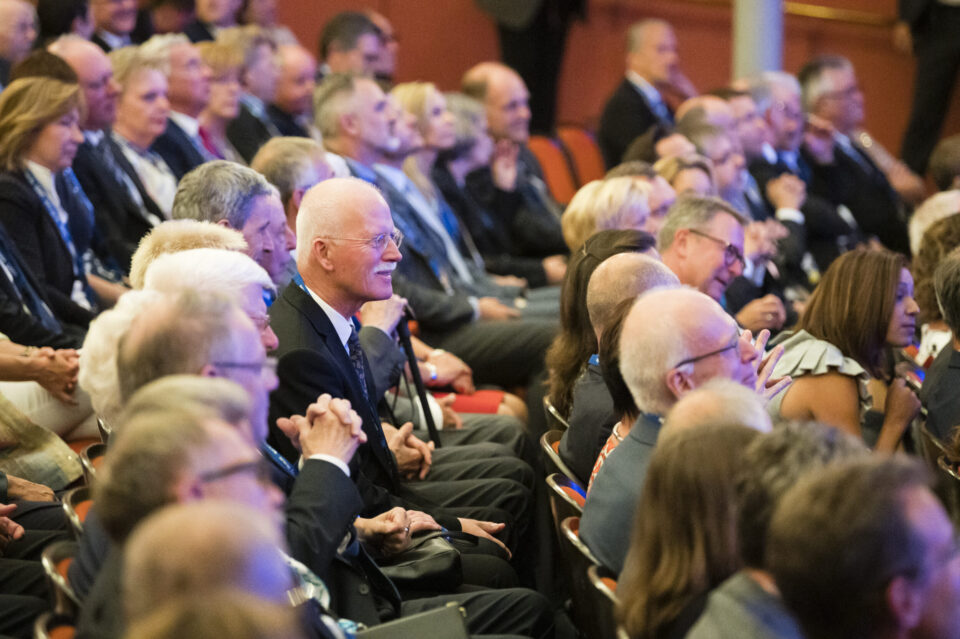Viele Personen sitzen in einem Auditorium oder Konferenzsaal in Reihen angeordneten Stühlen. Sie tragen formelle Kleidung und wirken aufmerksam, als würden sie einer Veranstaltung wie einer Konferenz oder einem Seminar folgen.