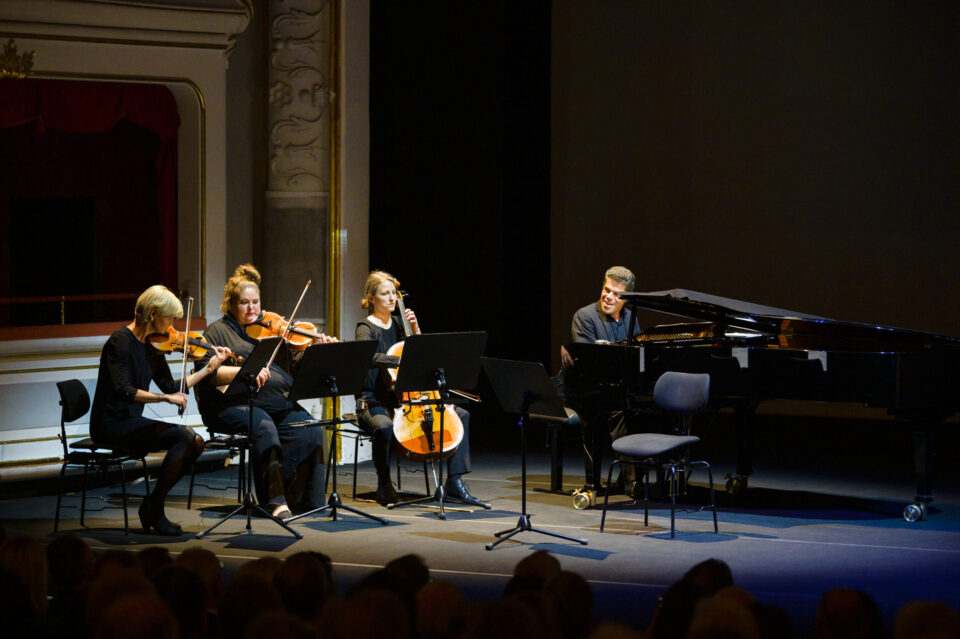 Vier Musikerinnen und Musiker treten auf einer Bühne in einem klassisch dekorierten Konzertsaal auf. Zu sehen sind zwei Violinistinnen oder Violinisten, eine Cellistin oder ein Cellist sowie eine Person am Klavier, alle mit Notenständern vor sich. Im unteren Bildbereich ist das Publikum zu erkennen, was auf ein Live-Konzert hinweist.